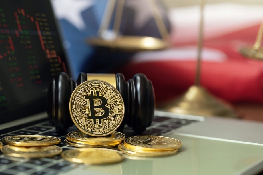 Eine Bitcoin-Münze liegt auf einem Richterhammer, umgeben von Krypto-Münzen. Im Hintergrund sind auf einem Laptop Aktiencharts und eine verschwommene amerikanische Flagge und Waagen zu sehen.