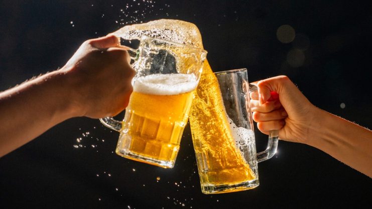 Zwei Hände stoßen mit Bierkrügen aneinander, sodass das Bier vor einem dunklen Hintergrund spritzt und schäumt – wir feiern Bitcoins Allzeithoch mit Freibier.