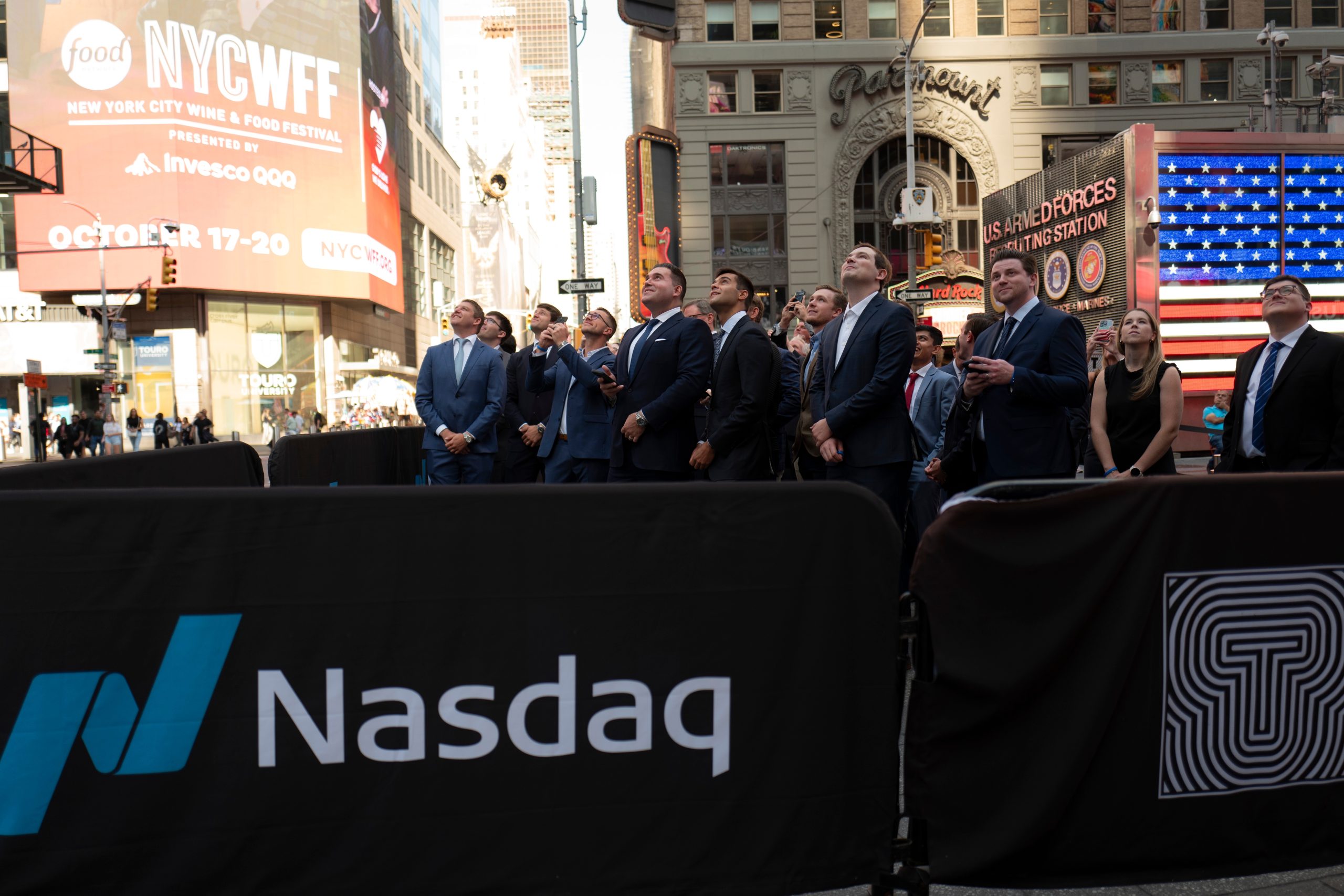 Eine Gruppe von Menschen in formeller Kleidung steht hinter einer Absperrung mit Nasdaq-Logo am Times Square und blickt nach oben. Digitale Werbetafeln zeigen das CoinShares-Logo, im Hintergrund ist eine amerikanische Flagge zu sehen.