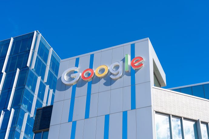 Das Google-Logo prangt deutlich auf der Außenseite eines modernen weißen Glasgebäudes vor einem klaren blauen Himmel.