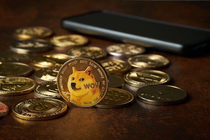Eine Dogecoin-Münze mit einem Shiba Inu steht aufrecht zwischen verschiedenen anderen Kryptowährungsmünzen und symbolisiert, wie Memecoin-Entwickler Geld verdienen können – und erinnert Investoren gleichzeitig daran, auf Rug Pulls zu achten – vor einem Smartphone auf einer braunen Oberfläche.