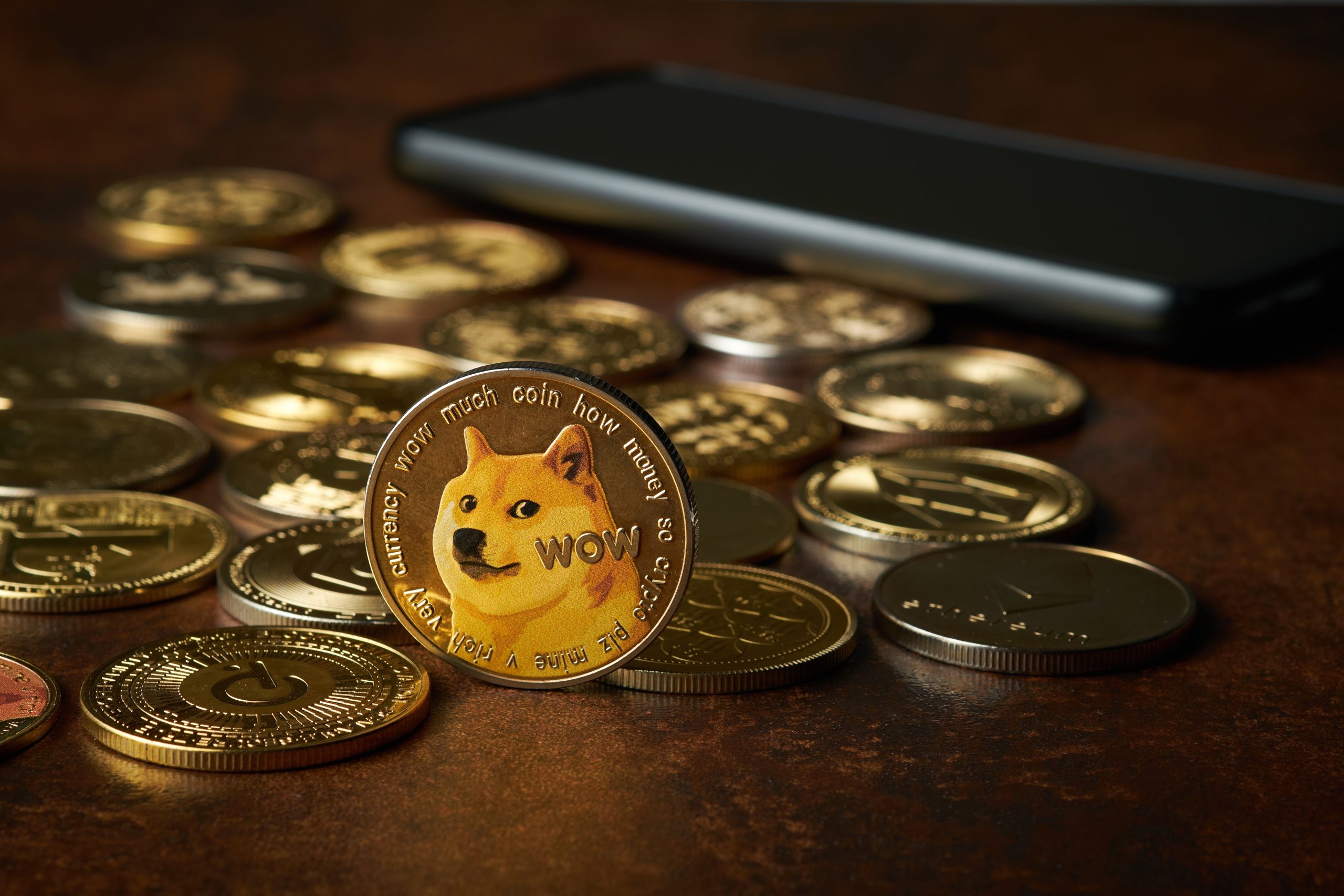 Eine Dogecoin-Münze mit einem Shiba Inu steht aufrecht zwischen verschiedenen anderen Kryptowährungsmünzen und symbolisiert, wie Memecoin-Entwickler Geld verdienen können – und erinnert Investoren gleichzeitig daran, auf Rug Pulls zu achten – vor einem Smartphone auf einer braunen Oberfläche.