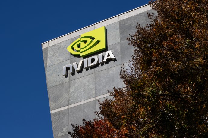 Das Logo und der Name von Nvidia sind prominent an der Außenseite eines modernen Gebäudes angebracht, mit Baumzweigen im Vordergrund und einem klaren blauen Himmel, der eine eindrucksvolle Kulisse bietet.