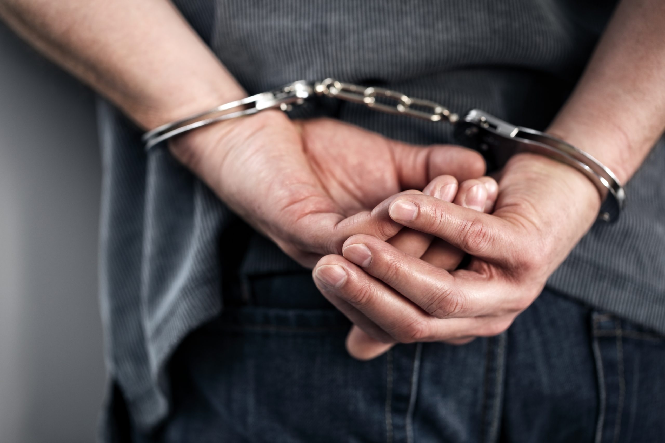 Eine Person in grauem Hemd und Jeans, von hinten gezeigt, mit in silbernen Handschellen gefesselten Händen, steht nach einem Krypto-bezogenen Vorfall vor der Festnahme.