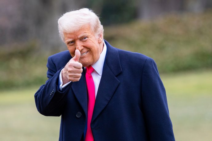Ein Mann in einem dunklen Mantel und einer leuchtend rosa Krawatte, die an Trumps typischen Stil erinnert, steht im Freien, zeigt den Daumen nach oben und lächelt.