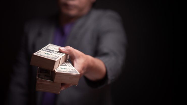 Eine Person im Anzug streckt eine Hand aus und hält drei Bündel Hundert-Dollar-Scheine in der Hand. Der unscharfe Hintergrund lässt auf eine Kryptobranche-Transaktion mit hohem Risiko schließen.