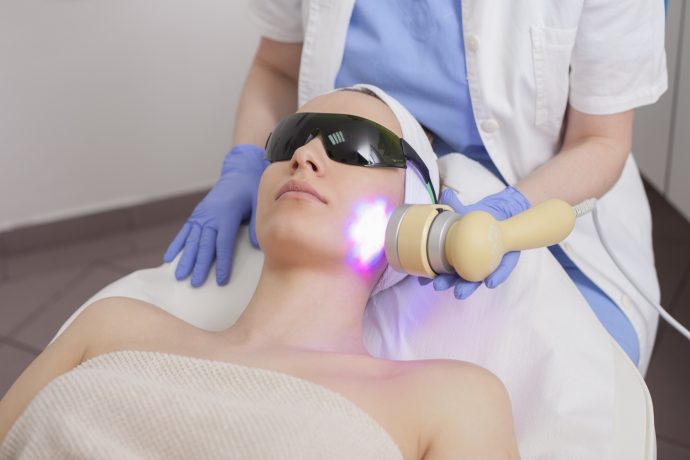 Eine Person mit Schutzbrille erhält in einer klinischen Umgebung von einem behandschuhten Fachmann eine Gesichtsbehandlung mit einem lichtemittierenden Krypto-Gerät.
