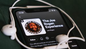 Auf einem Smartphone wird der Podcast „The Joe Rogan Experience“ auf Spotify angezeigt, in dem es um Bitcoin geht. Auf dem Gerät liegen vor einem dunkelgrünen Hintergrund weiße Kopfhörer.