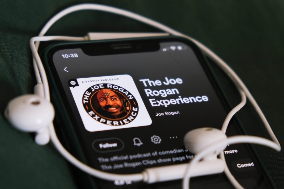 Auf einem Smartphone wird der Podcast „The Joe Rogan Experience“ auf Spotify angezeigt, in dem es um Bitcoin geht. Auf dem Gerät liegen vor einem dunkelgrünen Hintergrund weiße Kopfhörer.