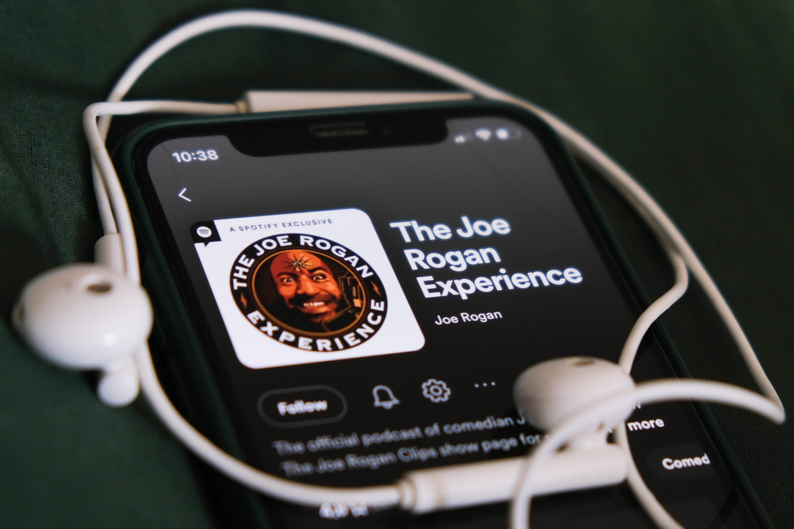 Auf einem Smartphone wird der Podcast „The Joe Rogan Experience“ auf Spotify angezeigt, in dem es um Bitcoin geht. Auf dem Gerät liegen vor einem dunkelgrünen Hintergrund weiße Kopfhörer.