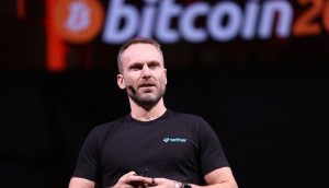 Paolo Ardoino in einem schwarzen Tether-T-Shirt spricht auf der Bühne, während auf dem Bildschirm hinter ihm ein großes Bitcoin-Logo und der Name Twenty One Capital angezeigt werden.