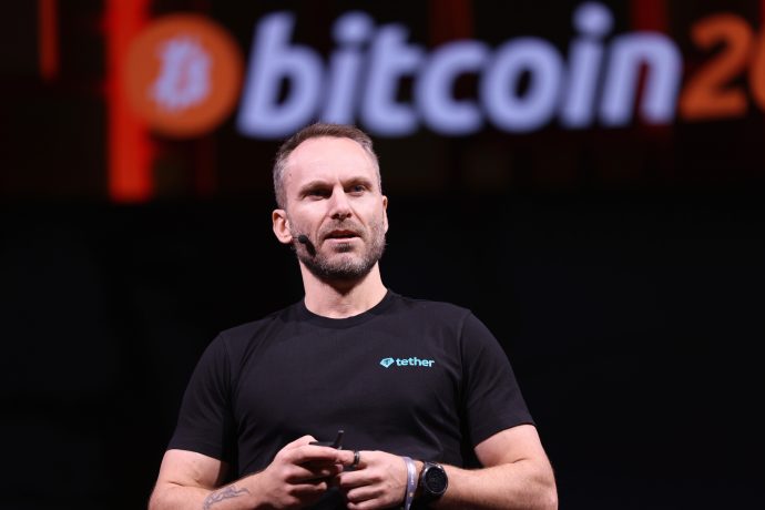 Paolo Ardoino in einem schwarzen Tether-T-Shirt spricht auf der Bühne, während auf dem Bildschirm hinter ihm ein großes Bitcoin-Logo und der Name Twenty One Capital angezeigt werden.