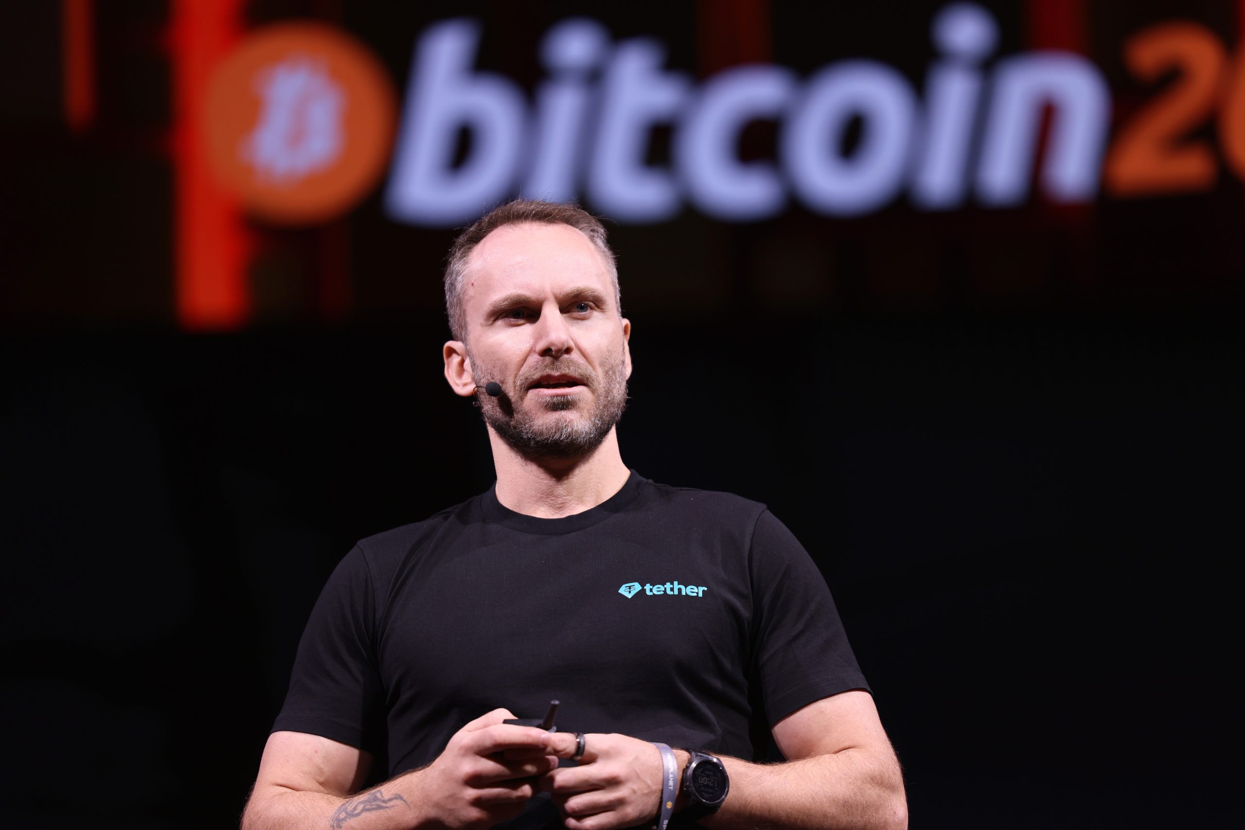Paolo Ardoino in einem schwarzen Tether-T-Shirt spricht auf der Bühne, während auf dem Bildschirm hinter ihm ein großes Bitcoin-Logo und der Name Twenty One Capital angezeigt werden.