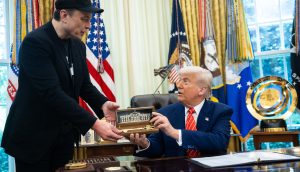 Ein Mann in schwarzem Outfit übergibt einem anderen Mann im Anzug, möglicherweise Trump, ein Modell des Weißen Hauses in einem Koffer. Der Mann sitzt an einem Schreibtisch in einem mit amerikanischen Flaggen und Militärabzeichen geschmückten Büro, was auf Diskussionen über den Krypto-Markt hindeutet.