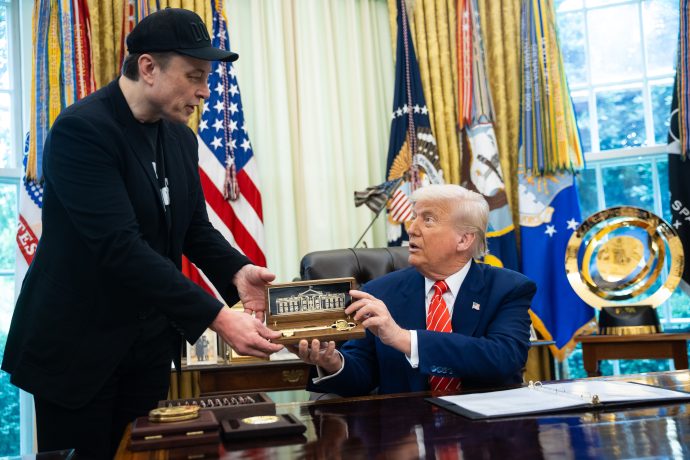 Ein Mann in schwarzem Outfit übergibt einem anderen Mann im Anzug, möglicherweise Trump, ein Modell des Weißen Hauses in einem Koffer. Der Mann sitzt an einem Schreibtisch in einem mit amerikanischen Flaggen und Militärabzeichen geschmückten Büro, was auf Diskussionen über den Krypto-Markt hindeutet.