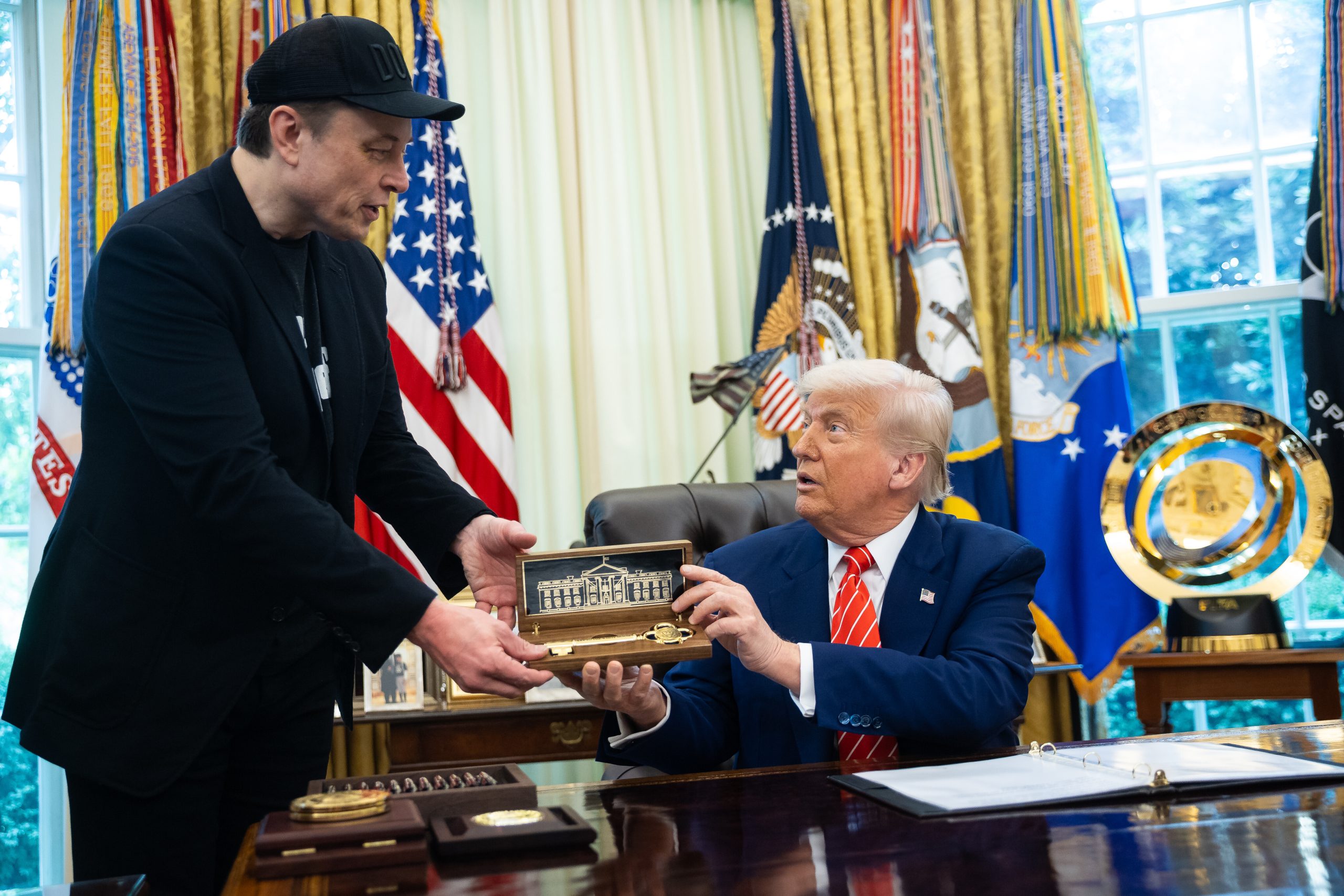 Ein Mann in schwarzem Outfit übergibt einem anderen Mann im Anzug, möglicherweise Trump, ein Modell des Weißen Hauses in einem Koffer. Der Mann sitzt an einem Schreibtisch in einem mit amerikanischen Flaggen und Militärabzeichen geschmückten Büro, was auf Diskussionen über den Krypto-Markt hindeutet.