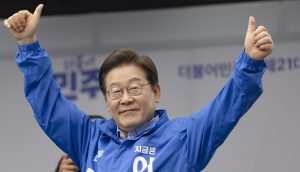 Lee Jae-myung mit Brille und blauer Jacke hebt beide Daumen hoch und lächelt. Er steht vor einem unscharfen Hintergrund mit koreanischem Text.