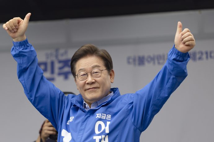 Lee Jae-myung mit Brille und blauer Jacke hebt beide Daumen hoch und lächelt. Er steht vor einem unscharfen Hintergrund mit koreanischem Text.