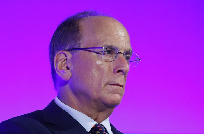 Ein Mann im Anzug und mit Brille wird vor einem lilafarbenen Hintergrund gezeigt, der leicht zur Seite blickt und damit an die souveräne Zuversicht erinnert, die man oft bei den Bitcoin-ETF-Präsentationen von BlackRock sieht.