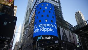 Eine blaue digitale Werbetafel auf einem städtischen Gebäude zeigt die Botschaft "It's happening with Ripple" über einem Nasdaq-Logo und erregt damit Aufmerksamkeit inmitten eines belebten Stadtgebiets, in dem sich der Prozess und die Berufung von Ripple entfalten.