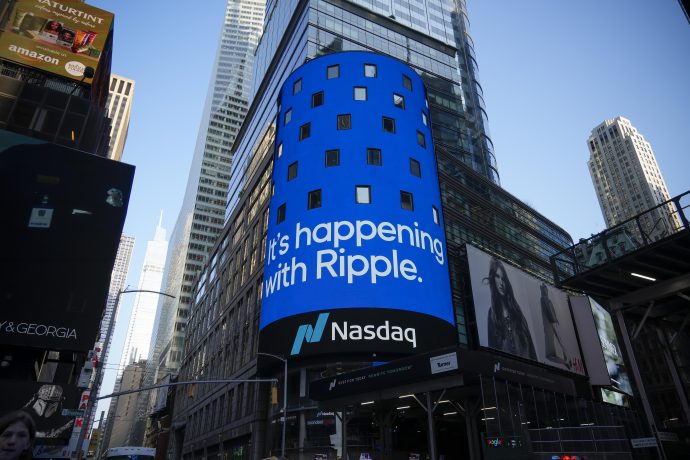 Eine blaue digitale Werbetafel auf einem städtischen Gebäude zeigt die Botschaft "It's happening with Ripple" über einem Nasdaq-Logo und erregt damit Aufmerksamkeit inmitten eines belebten Stadtgebiets, in dem sich der Prozess und die Berufung von Ripple entfalten.