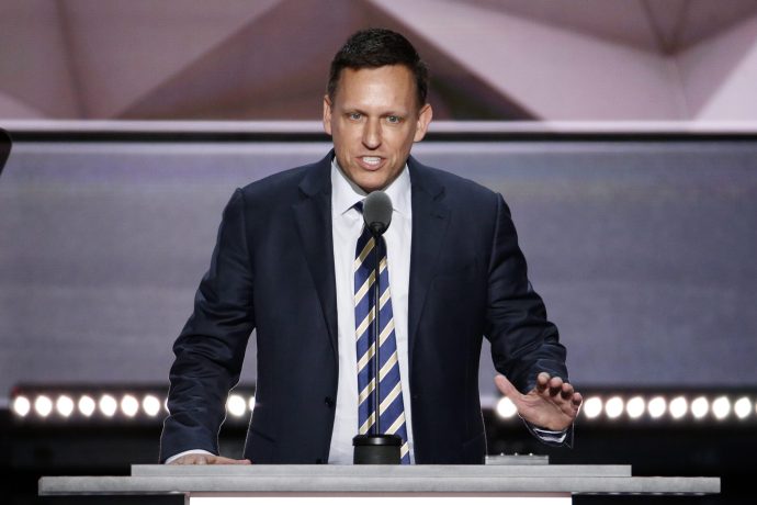 Peter Thiel in Anzug und gestreifter Krawatte spricht an einem Podium mit Mikrofon und gestikuliert mit einer Hand vor einem geometrischen Hintergrund.
