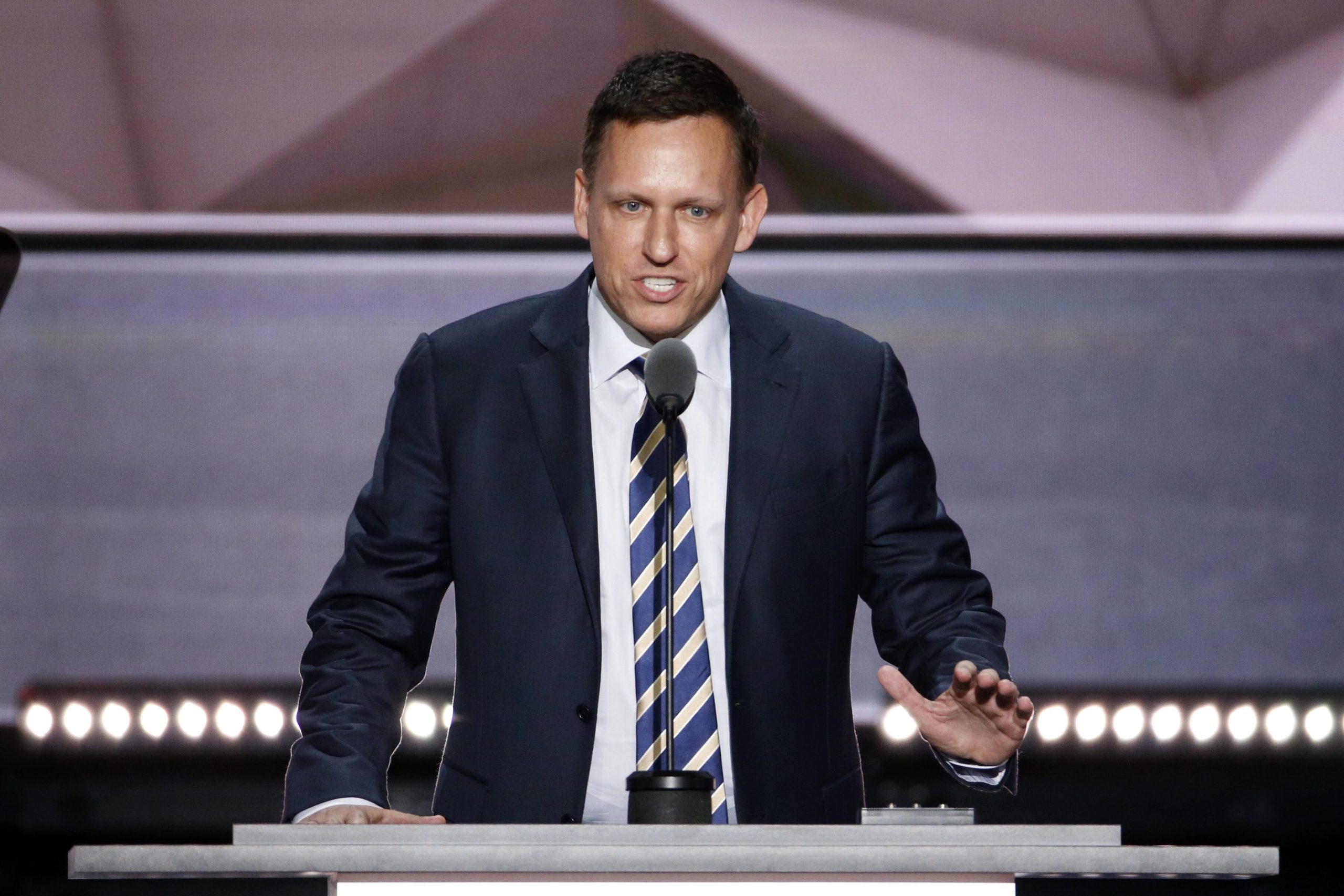 Peter Thiel in Anzug und gestreifter Krawatte spricht an einem Podium mit Mikrofon und gestikuliert mit einer Hand vor einem geometrischen Hintergrund.
