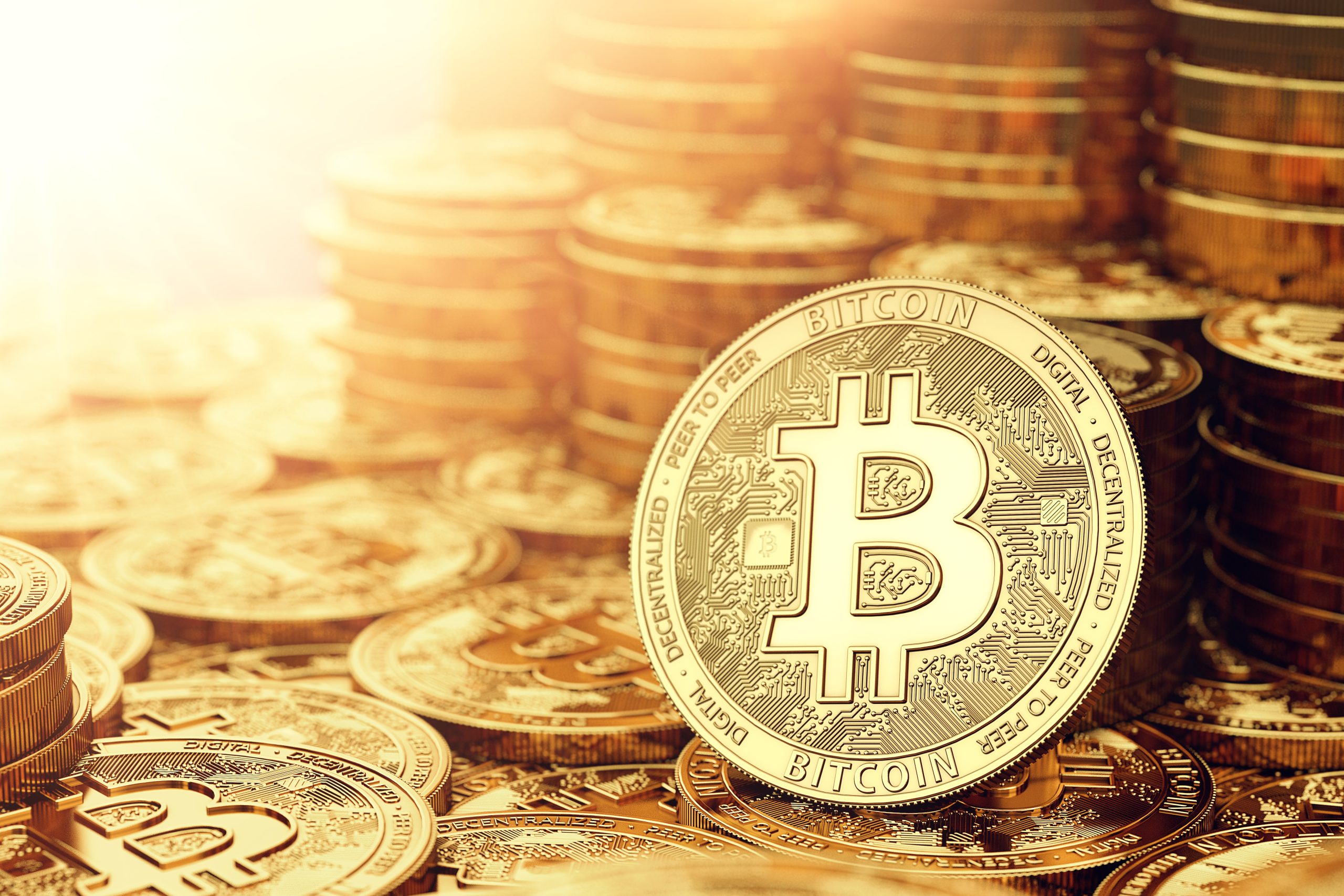 Nahaufnahme mehrerer goldfarbener Bitcoin-Münzen, wobei eine im Vordergrund aufrecht steht und das Sonnenlicht aus der oberen linken Ecke scheint.