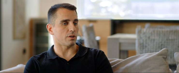 Ein Bitcoin-Influencer in einem schwarzen Polohemd sitzt auf einer Couch im Haus und schaut mit neutraler Miene nach links.