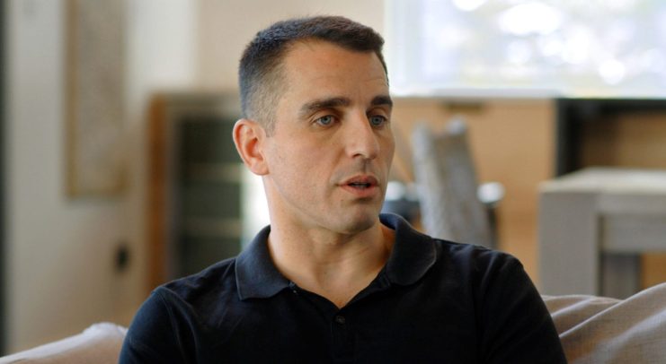 Ein Bitcoin-Influencer in einem schwarzen Polohemd sitzt auf einer Couch im Haus und schaut mit neutraler Miene nach links.