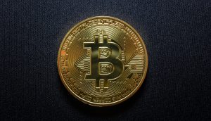 Eine goldfarbene physische Bitcoin-Münze mit dem Bitcoin-Symbol ist auf einer schwarz strukturierten Oberfläche platziert und erinnert an die Aufregung während des Bitcoin-Allzeithochs, das von Krypto-Führern wie CZ und Arthur Hayes gefeiert wurde.