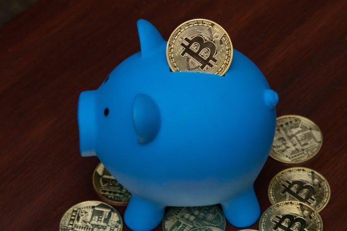 Ein blaues Sparschwein mit Bitcoin-Münzen im Inneren und rundherum auf einer hölzernen Oberfläche, perfekt um die Idee eines Bitcoin-Sparplans sinnvoll zu illustrieren.