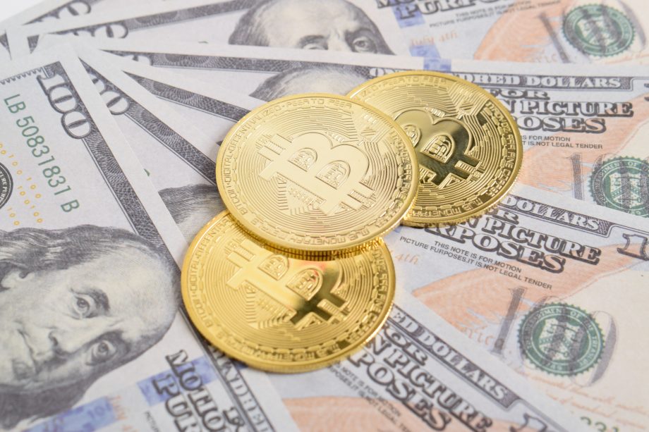 Drei goldfarbene Bitcoin-Münzen ruhen auf mehreren ausgebreiteten US-Hundert-Dollar-Scheinen und verdeutlichen die Überschneidung von Bitcoin und herkömmlicher Währung.