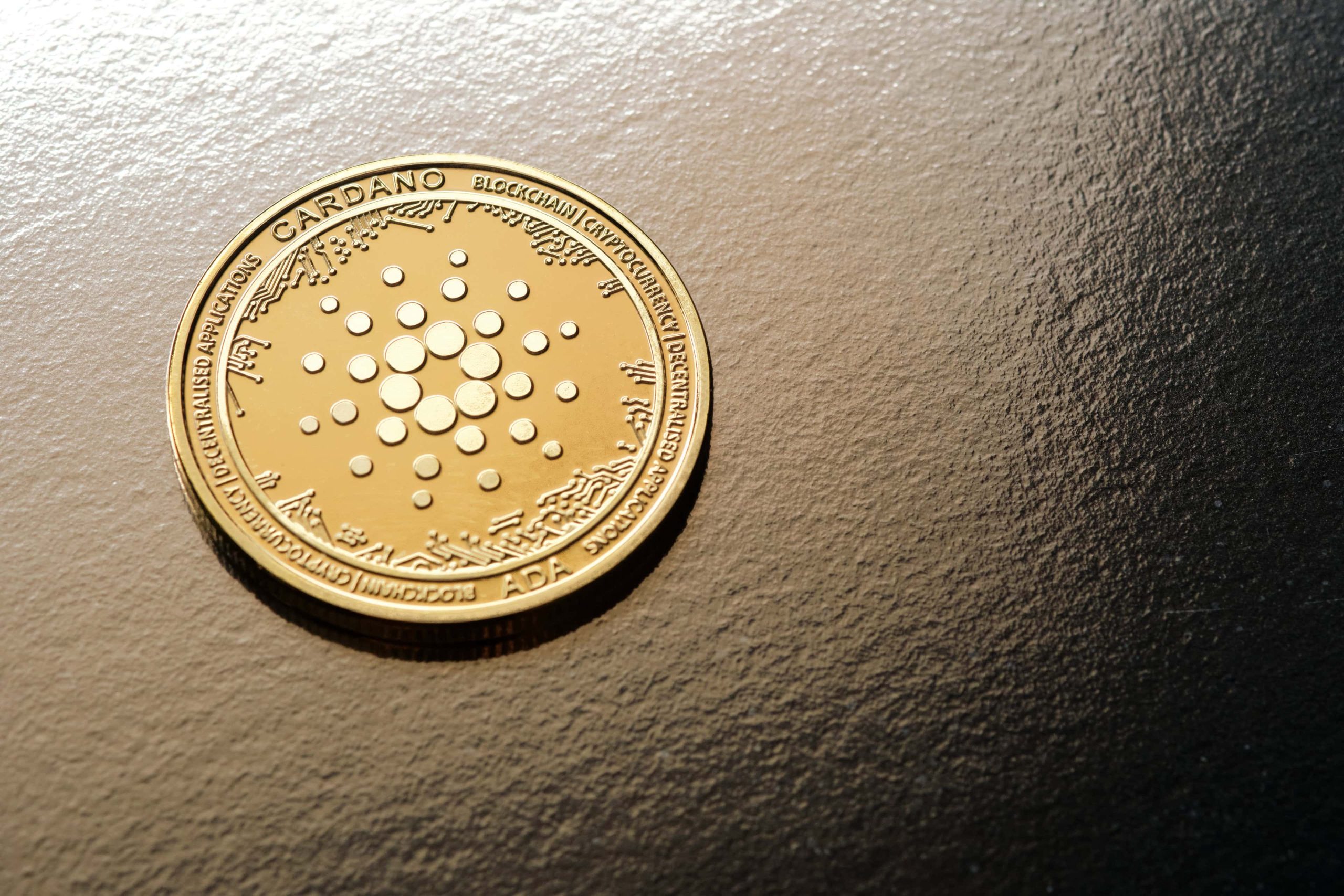 Eine goldfarbene Cardano (ADA) Kryptowährungsmünze ruht auf einer strukturierten, dunklen Oberfläche und zeigt das unverwechselbare Cardano-Logo und detaillierte Inschriften.