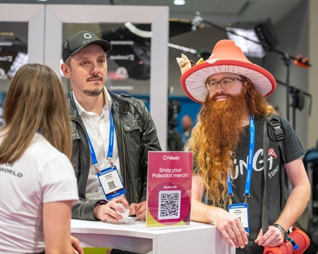 Drei Personen unterhalten sich an einem Stand mit einem Polkadot-Warenschild, einem QR-Code und einer Getränkestation; ein Mann trägt einen großen orangefarbenen Hut mit Plüschpilzen, während ein anderer eine schwarze Mütze und Jacke trägt.