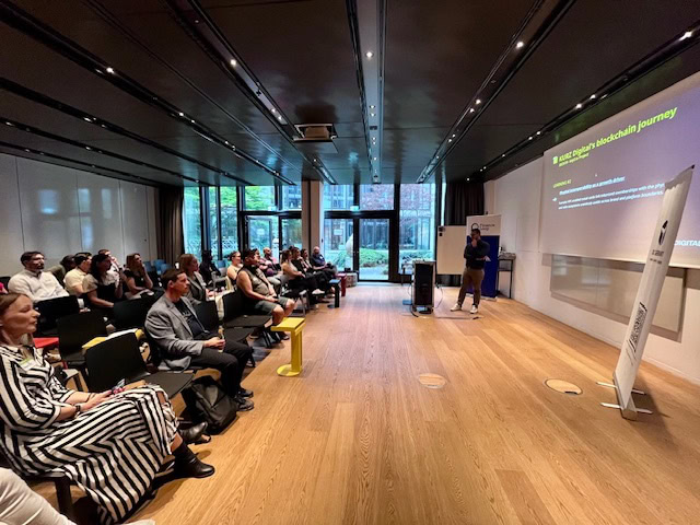 Eine Person präsentiert einem Publikum in einem modernen Konferenzraum mit großen Fenstern und Holzböden eine Diashow zum Thema Blockchain.