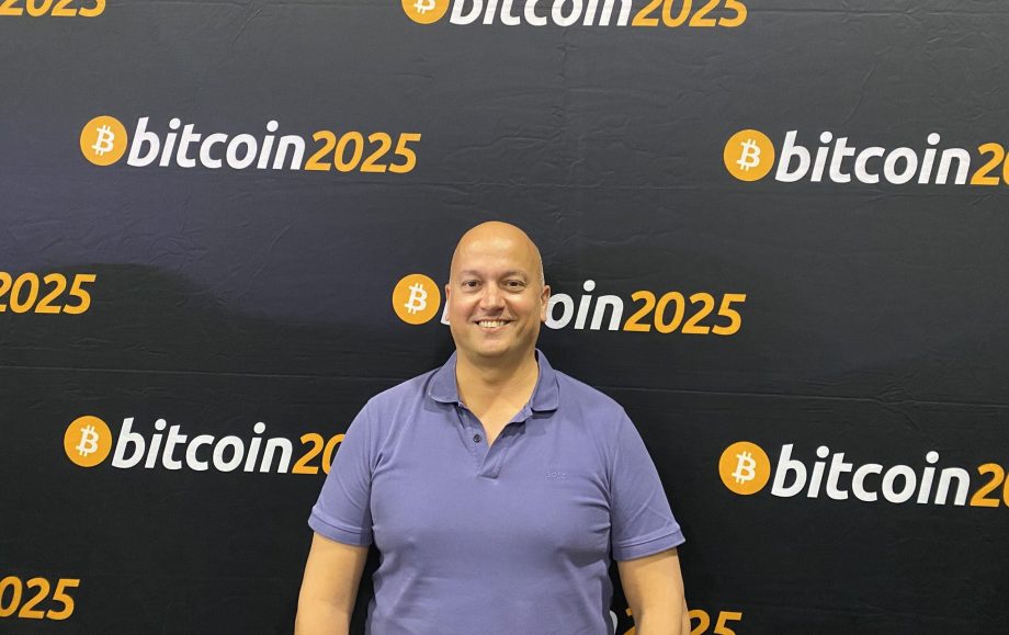Eine Person in einem lila Poloshirt steht vor einem Hintergrund, auf dem wiederholt der Text bitcoin2025 und das Bitcoin-Logo angezeigt werden.