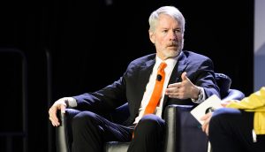 Michael Saylor, der Gründer von MicroStrategy, in Anzug und orangefarbener Krawatte sitzt auf einem Stuhl und gestikuliert mit seiner rechten Hand, während er während einer Podiumsdiskussion über die Bitcoin-Strategie spricht.