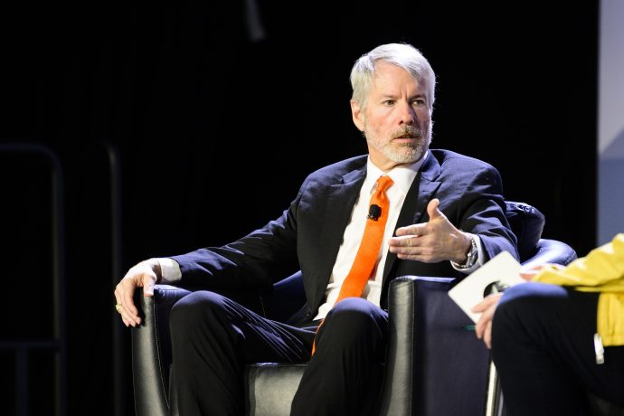 Michael Saylor, der Gründer von MicroStrategy, in Anzug und orangefarbener Krawatte sitzt auf einem Stuhl und gestikuliert mit seiner rechten Hand, während er während einer Podiumsdiskussion über die Bitcoin-Strategie spricht.