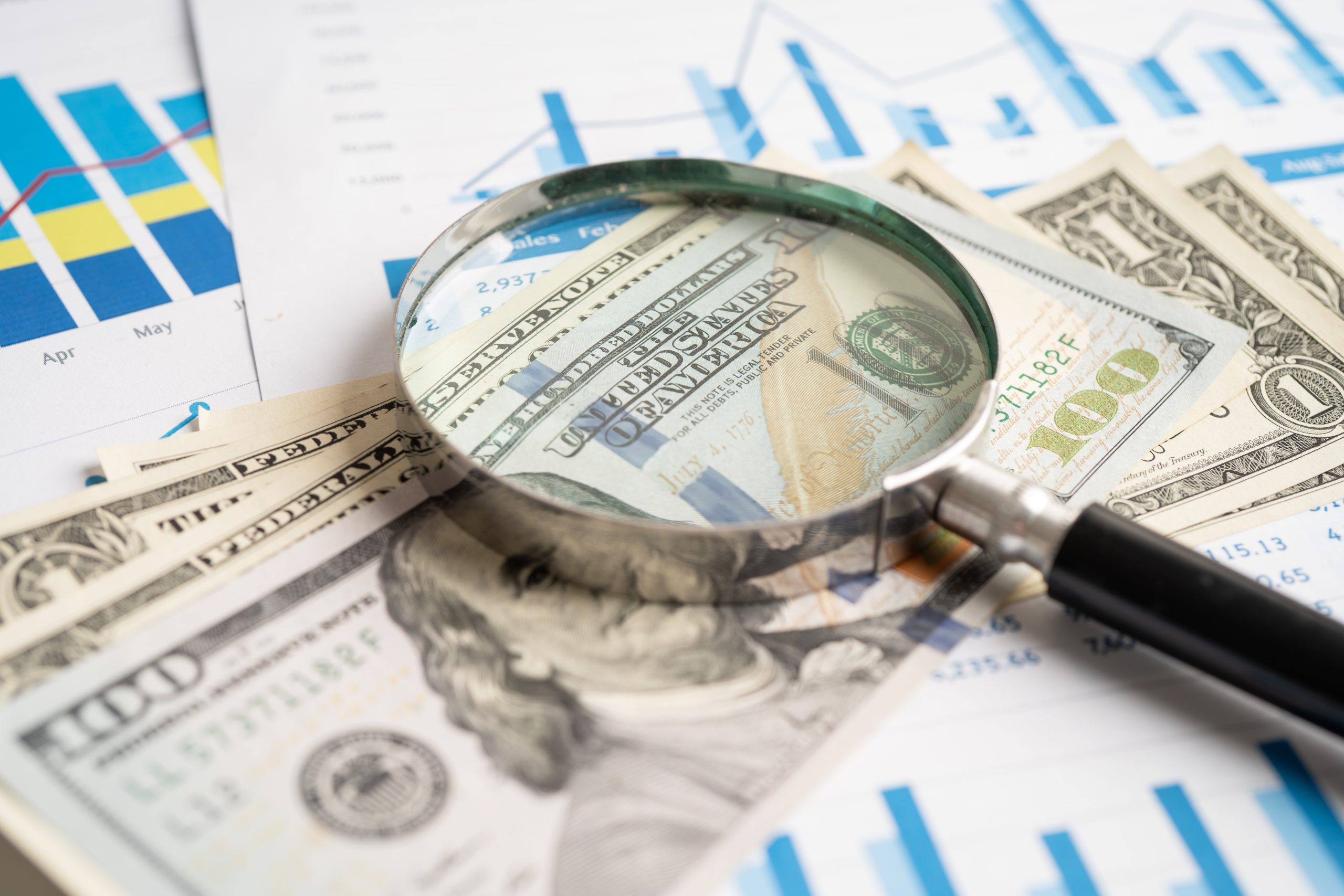Lupe über US-Dollar-Noten auf Finanztabellen und -grafiken, die die Finanzanalyse und die Untersuchung von Geld- oder Krypto-Daten darstellen.