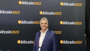 Ein Mann in einem blauen Anzug steht bei einer Veranstaltung zum Thema Bitcoin vor einem Hintergrund mit wiederholten Bitcoin 2025-Logos.