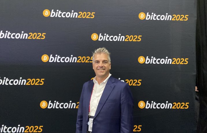 Ein Mann in einem blauen Anzug steht bei einer Veranstaltung zum Thema Bitcoin vor einem Hintergrund mit wiederholten Bitcoin 2025-Logos.