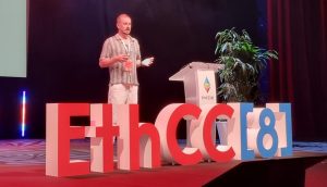 Thomas Stanczak steht auf der EthCC[8]-Konferenz auf der Bühne und hält einen Vortrag über Ethereum, während im Hintergrund eine Folie projiziert wird und die Bühne beleuchtet ist.