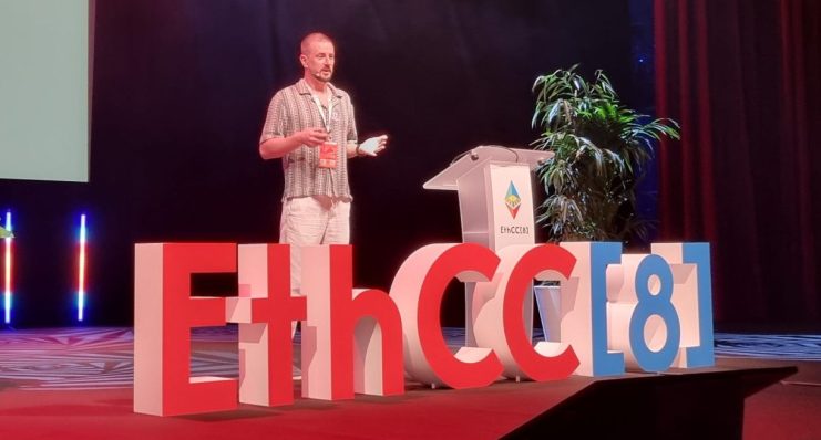Thomas Stanczak steht auf der EthCC[8]-Konferenz auf der Bühne und hält einen Vortrag über Ethereum, während im Hintergrund eine Folie projiziert wird und die Bühne beleuchtet ist.