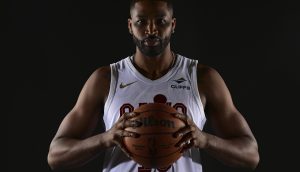 NBA-Star Tristan Thomas, in einem weißen Trikot, hält mit beiden Händen einen Basketball vor einem dunklen Hintergrund, bereit für das Interview.