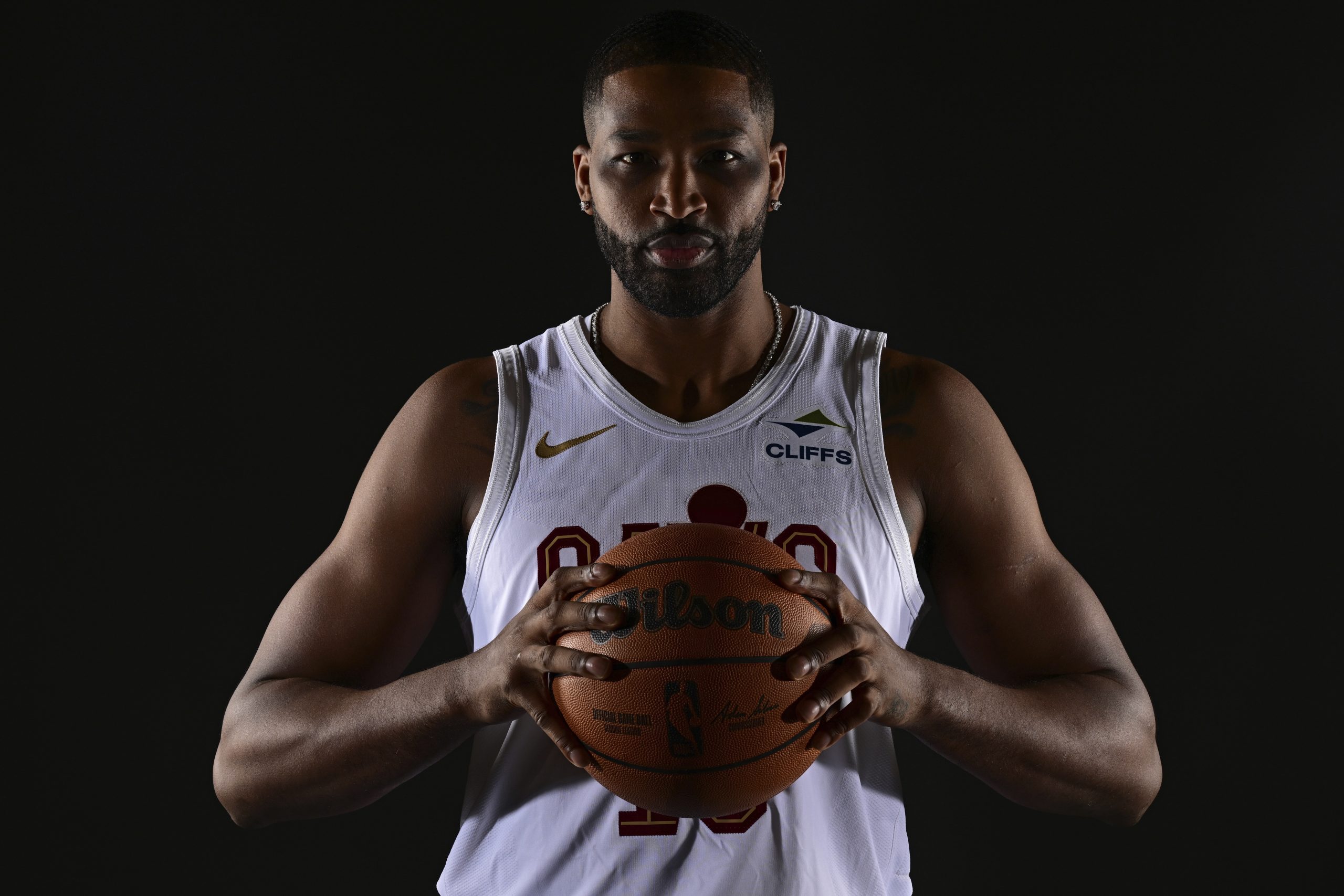 NBA-Star Tristan Thomas, in einem weißen Trikot, hält mit beiden Händen einen Basketball vor einem dunklen Hintergrund, bereit für das Interview.