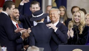 Donald Trump im Anzug umarmt seine Frau Melania mit Hut auf einem Podium, umgeben von seinen Kindern.