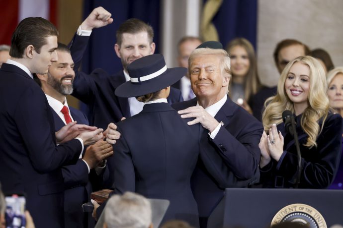 Donald Trump im Anzug umarmt seine Frau Melania mit Hut auf einem Podium, umgeben von seinen Kindern.