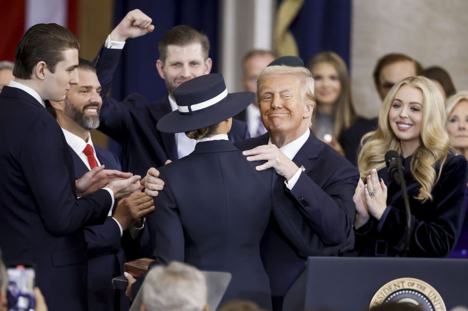 Donald Trump im Anzug umarmt seine Frau Melania mit Hut auf einem Podium, umgeben von seinen Kindern.