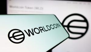 Auf dem Smartphone werden das Worldcoin-Logo und der Name angezeigt, was auf die Orb-Verifikation hindeutet, mit einer unscharfen größeren Version desselben Logos und Texts im Hintergrund.
